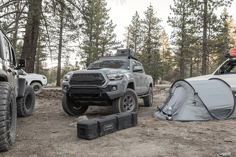 Load image into Gallery viewer, A truck in the forest with one Pelican BX85S case attached to a roof rack on it, and another BX85S case on the ground in front of it.
