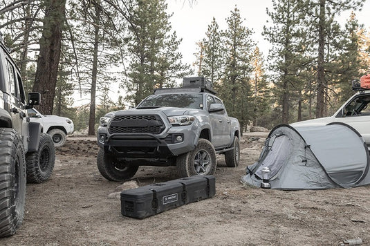 A truck in the forest with one Pelican BX85S case attached to a roof rack on it, and another BX85S case on the ground in front of it.